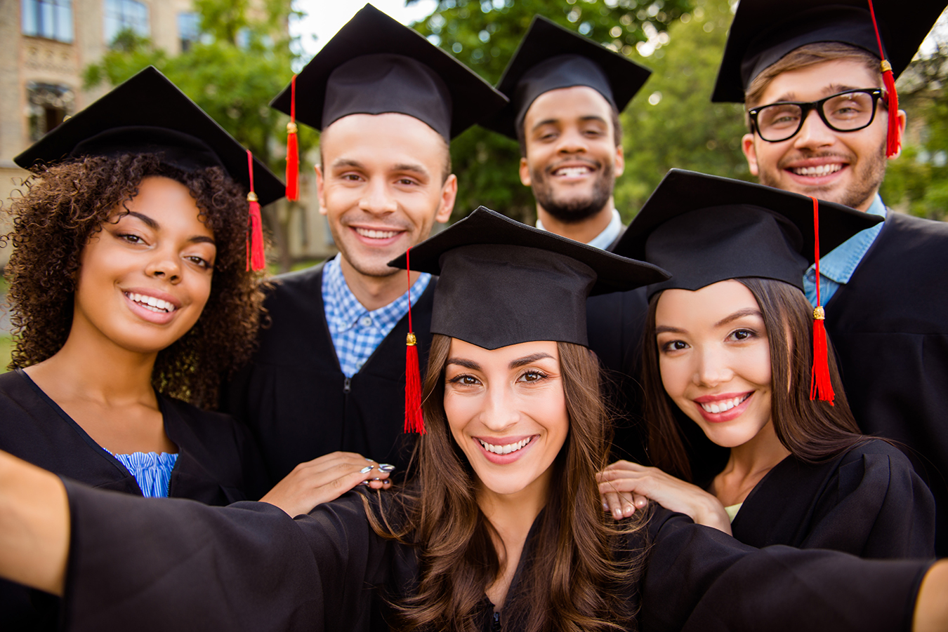 Selfie for memories. Six with cheerful graduates are posing for selfie shot, attractive brunette lady is taking, wearing gowns and mortar boards, outside on a summer day